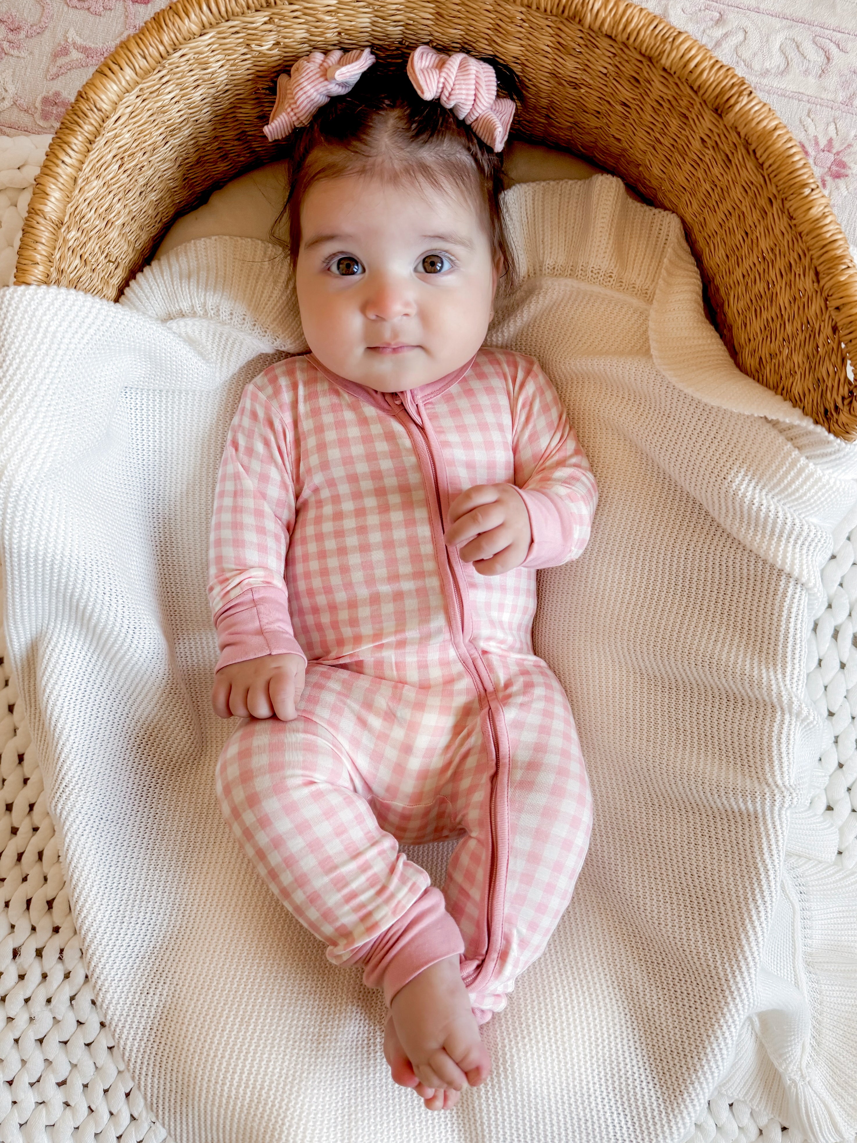 Baby in a pink checkered onesie sitting in a woven basket, surrounded by a cozy blanket.