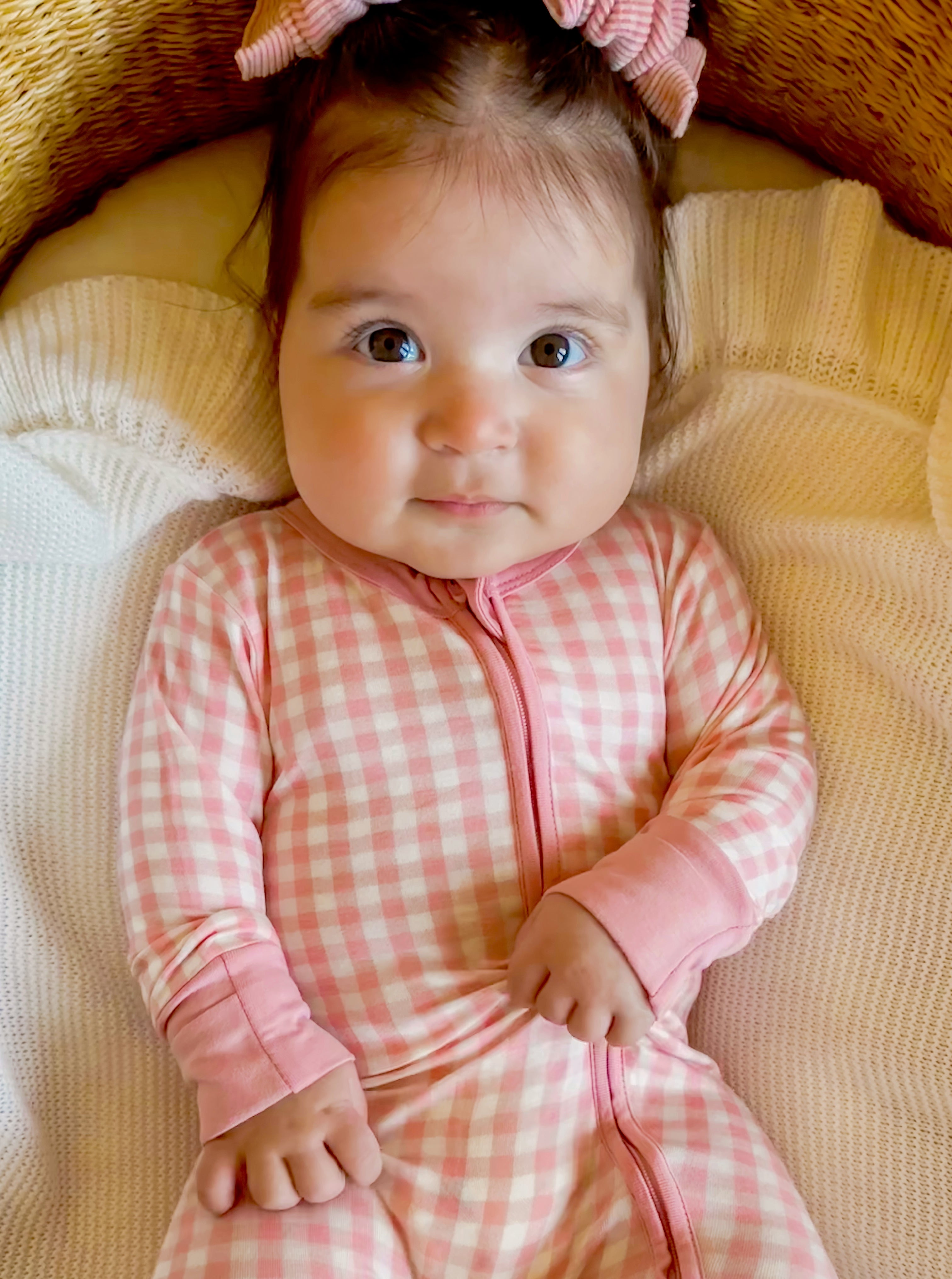 Infant with a pink checkered onesie and a bow, sitting on soft blankets, looking directly at the camera.