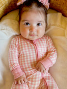 Infant with a pink checkered onesie and a bow, sitting on soft blankets, looking directly at the camera.
