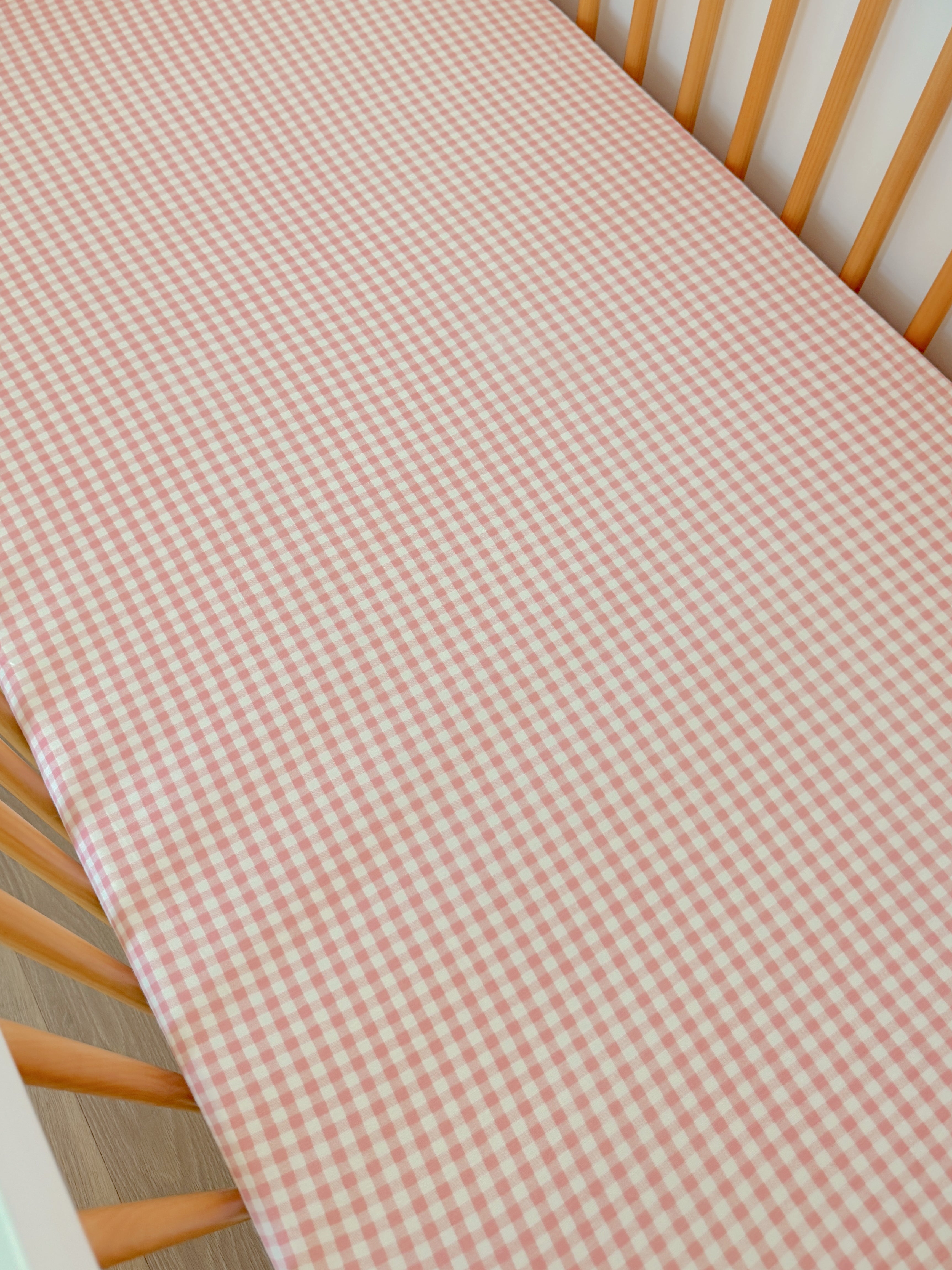 Pink and white gingham patterned crib sheet on a wooden crib.