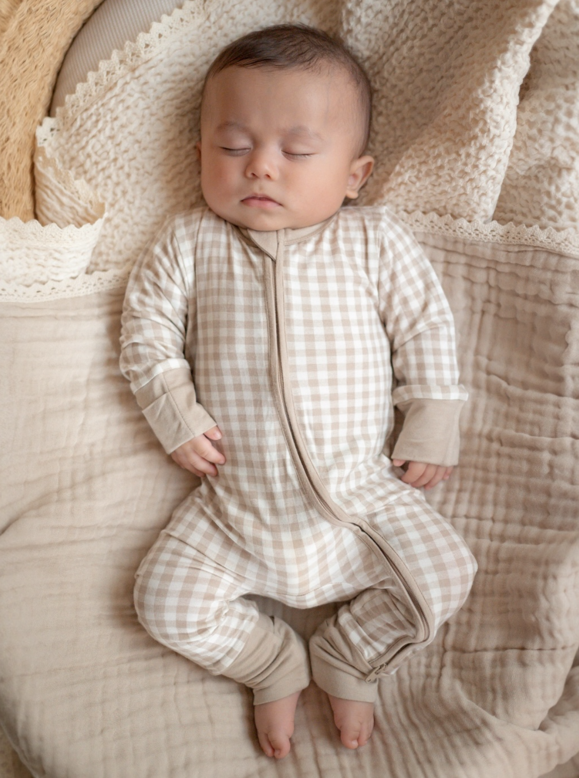 Sleepy baby in a beige checkered onesie, resting on soft, neutral-toned bedding.