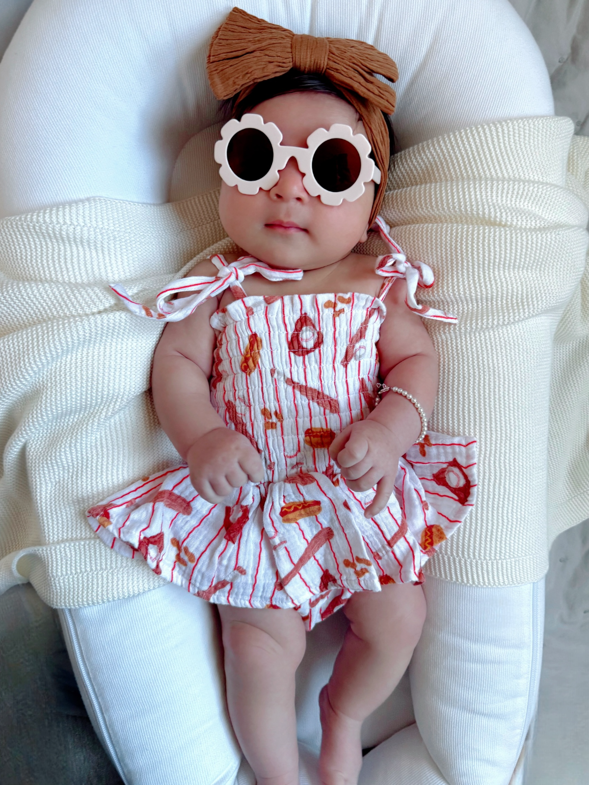 Baby wearing a floral headband and oversized sunglasses, lying on a white blanket, smiling at the camera.