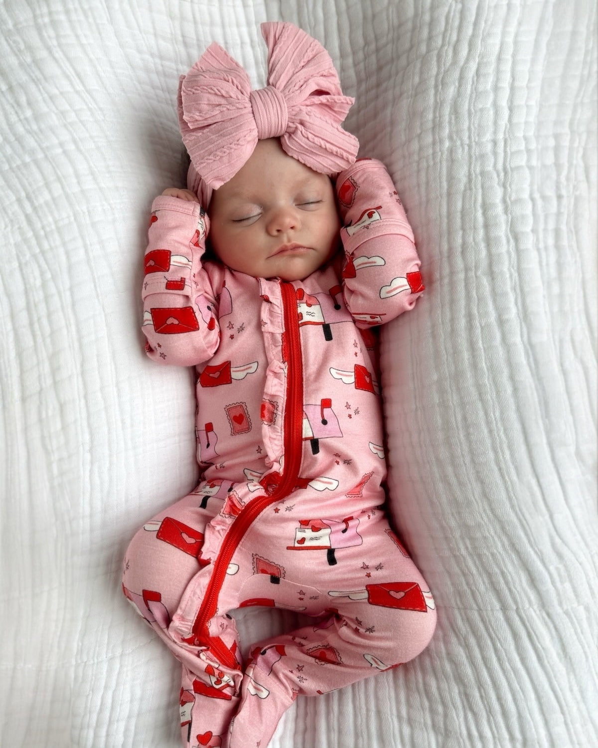 Infant in pink pajamas with red patterns and a large pink bow, peacefully sleeping on a white textured blanket.