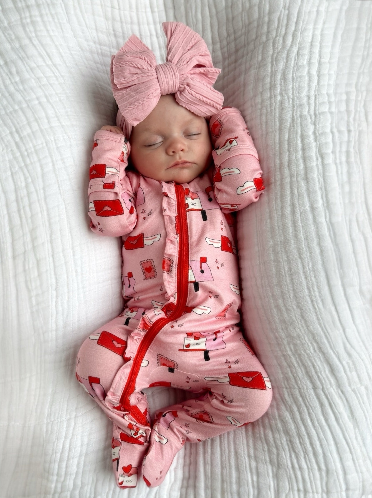 Infant in pink pajamas with red patterns and a large pink bow, peacefully sleeping on a white textured blanket.