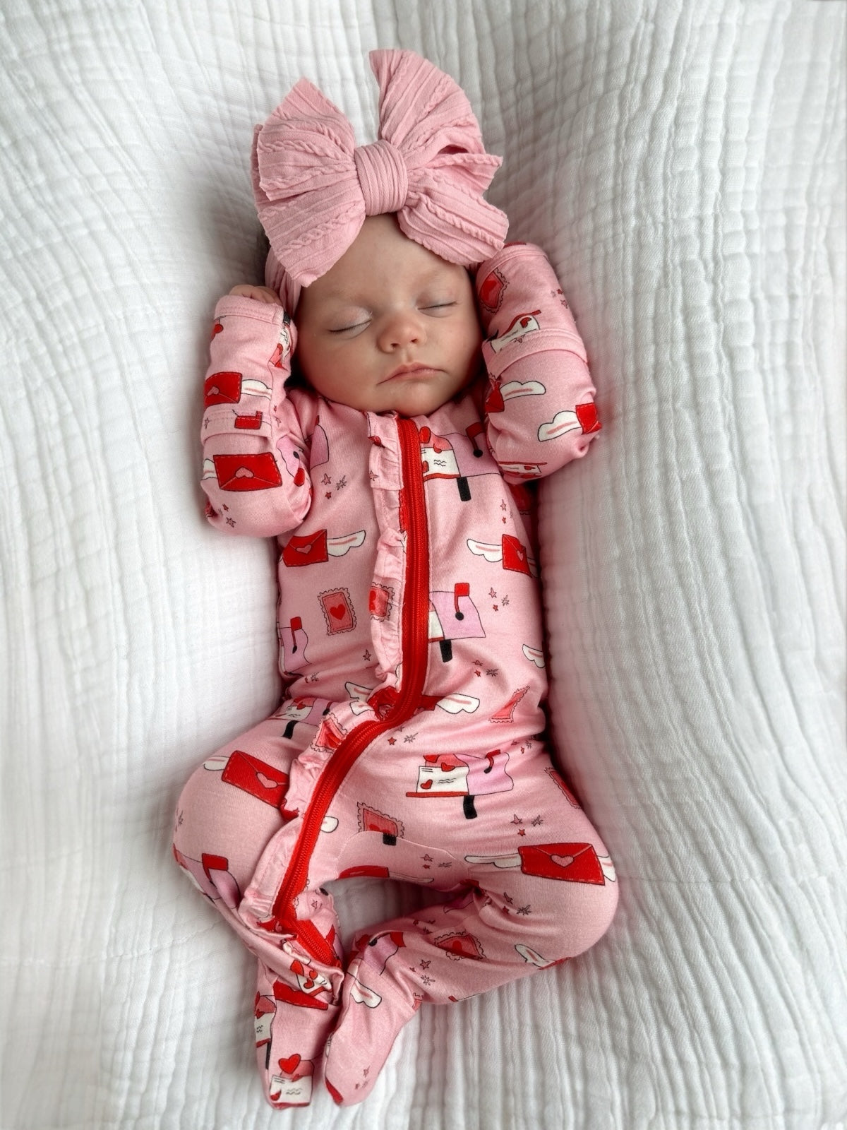 Infant in pink pajamas with red patterns and a large pink bow, peacefully sleeping on a white textured blanket.