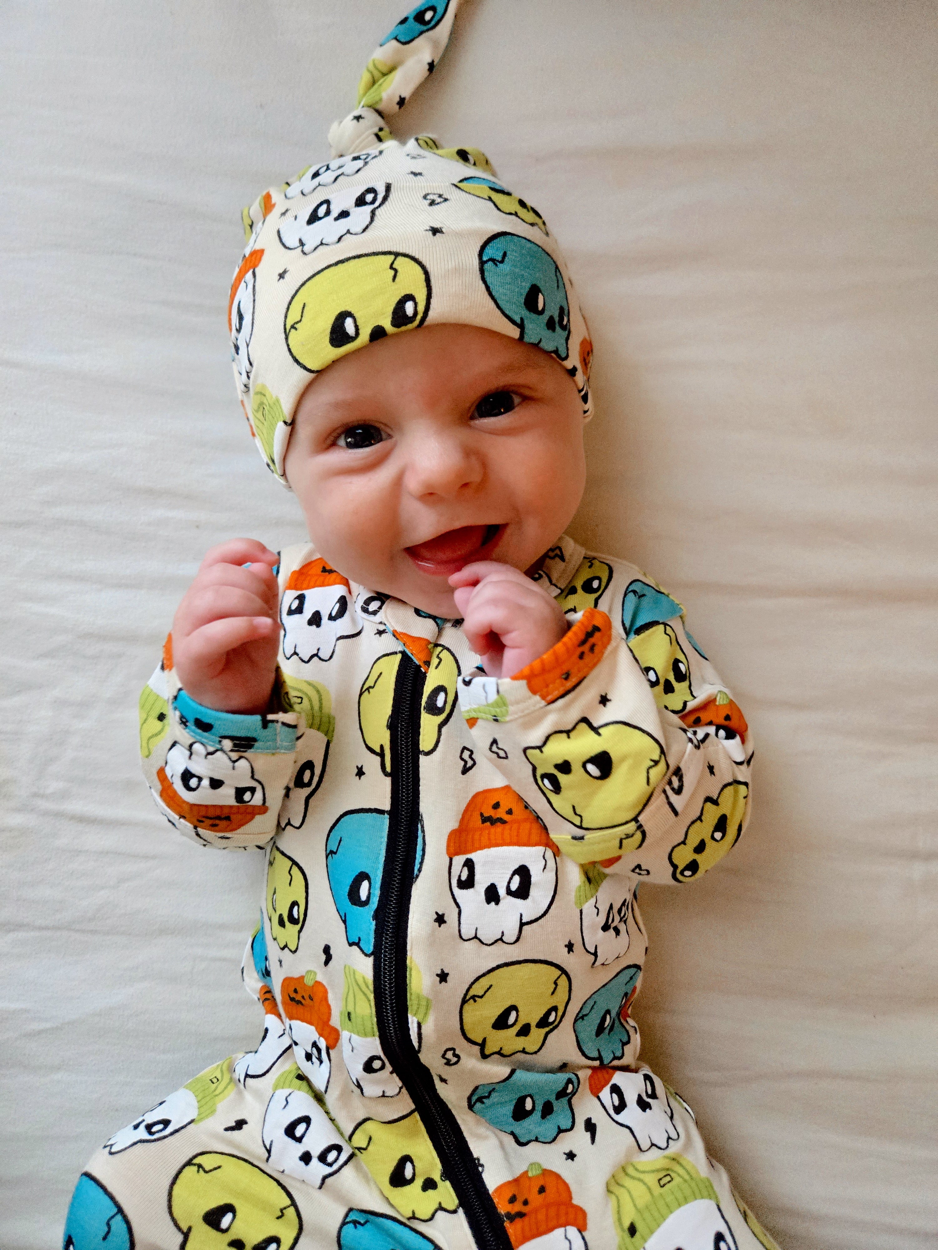 Smiling baby in colorful skull-patterned outfit and hat, sitting on a light-colored surface.