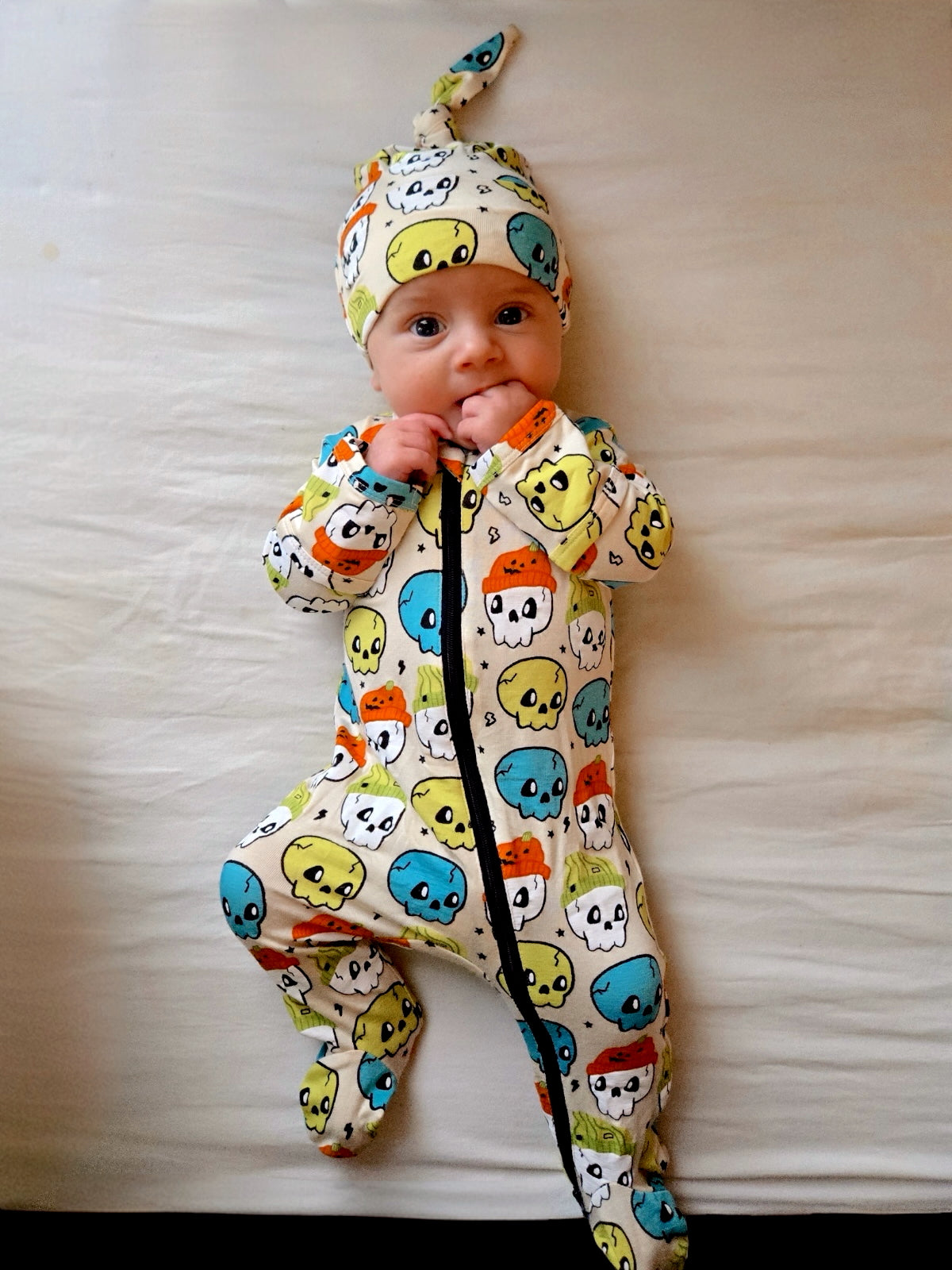 Infant wearing a colorful skull-patterned onesie and matching hat, lying on a light-colored surface.