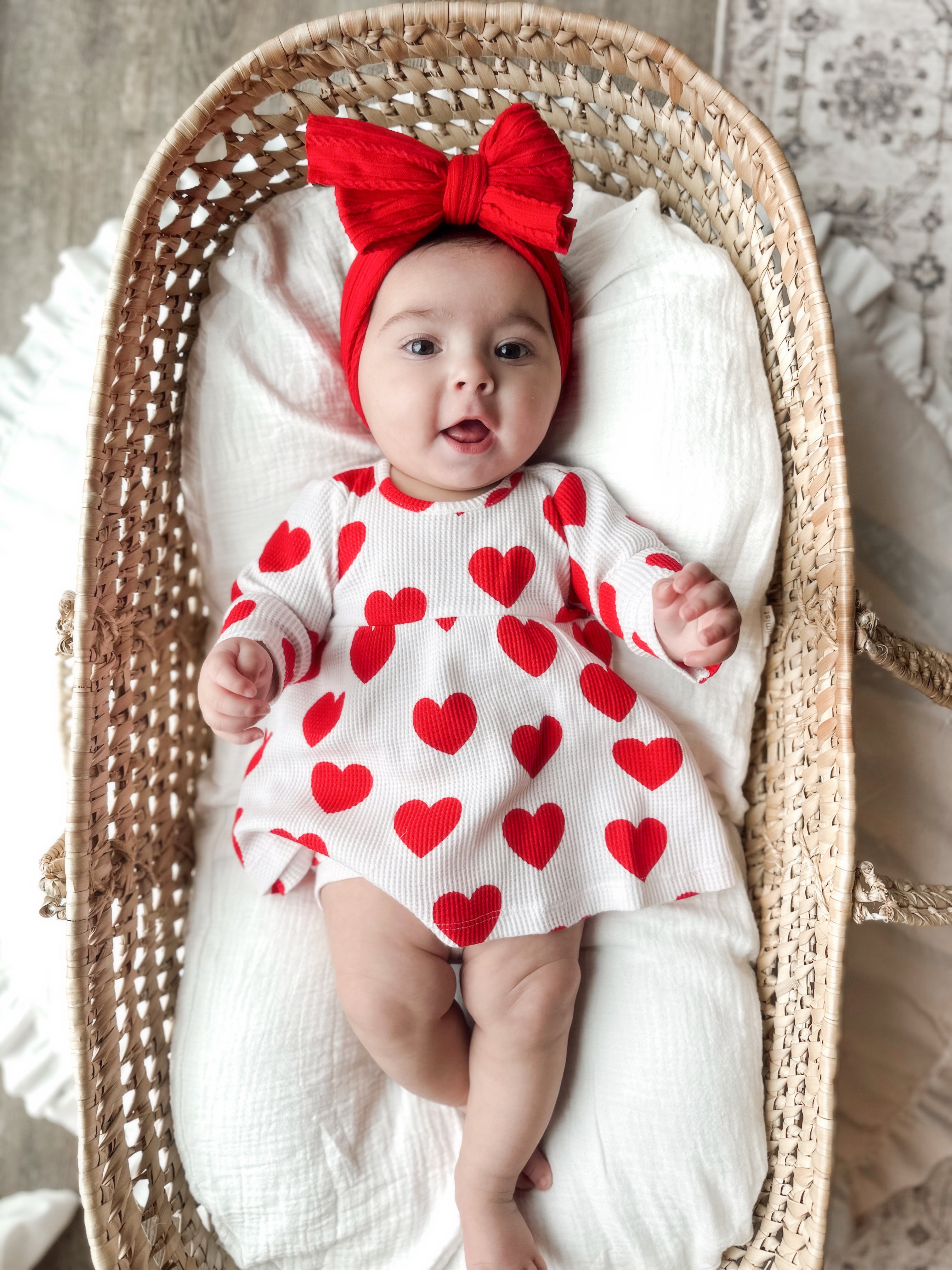 Smiling baby in a red heart-patterned dress and red headband, lying in a woven bassinet with a white blanket.