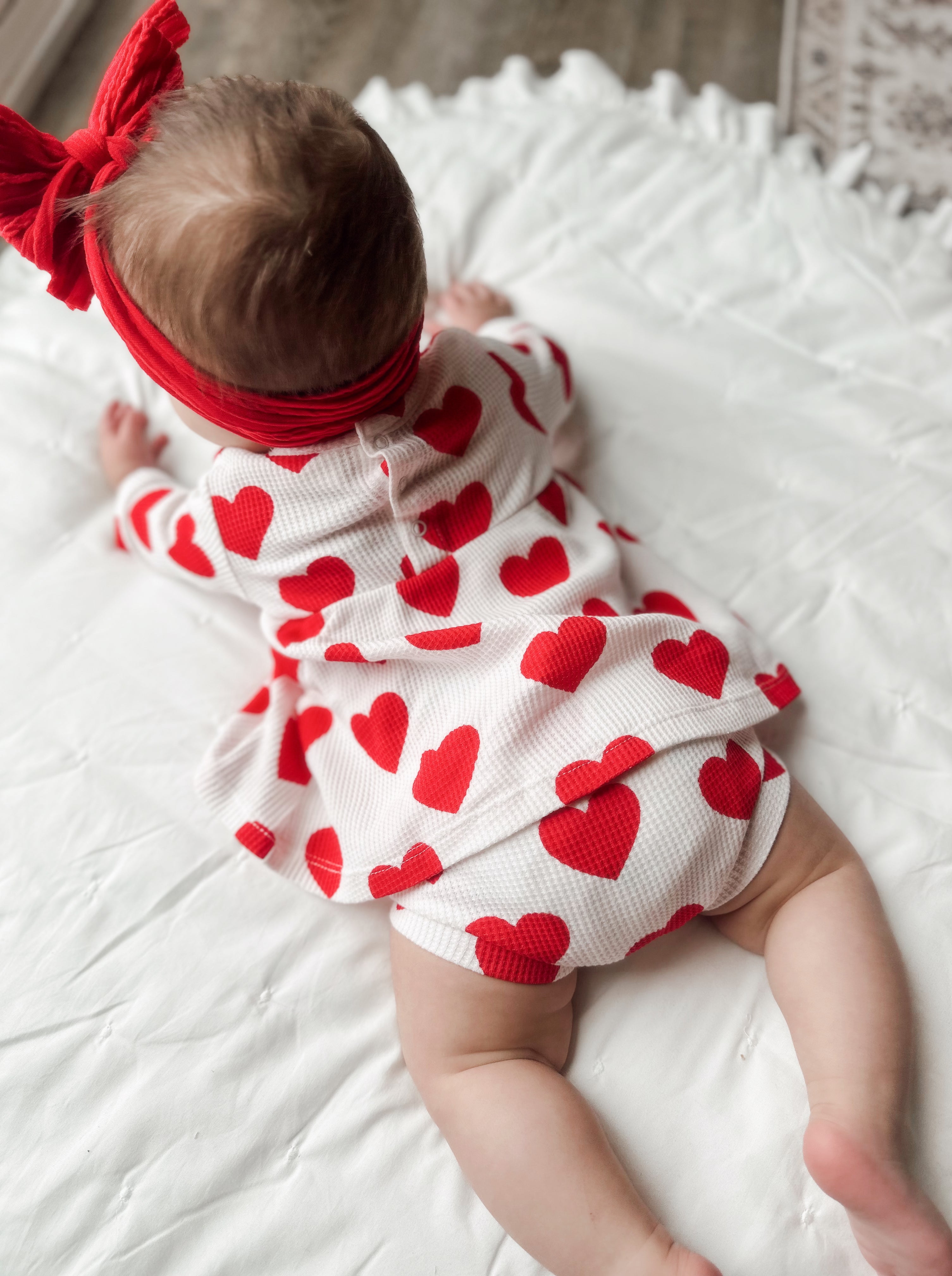 Baby girl in a heart-patterned outfit and red headband, crawling on a white textured blanket.