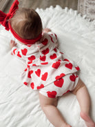 Baby girl in a heart-patterned outfit and red headband, crawling on a white textured blanket.