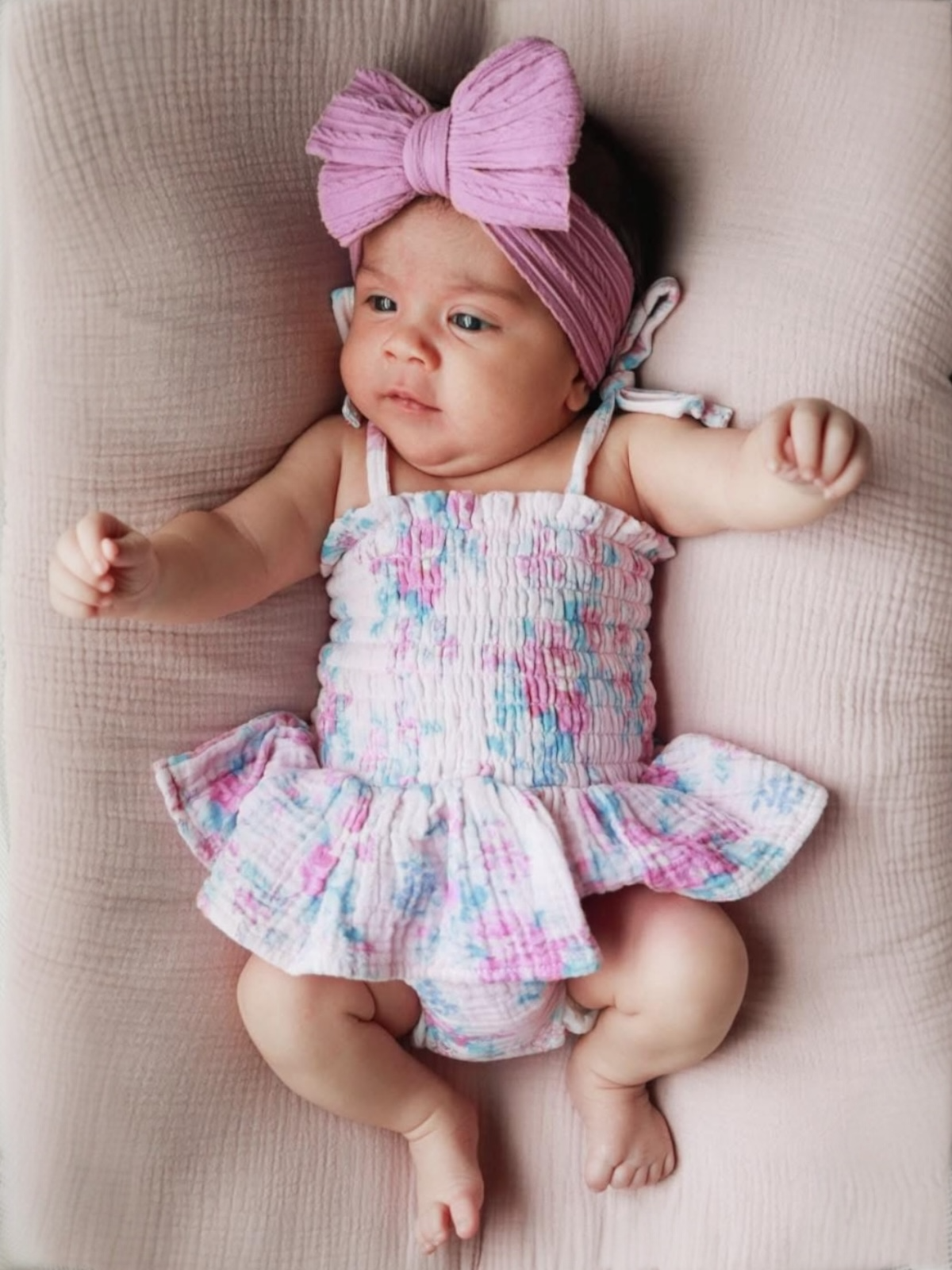Baby girl in a floral outfit with a pink bow, lying on a soft blanket, looking curious.