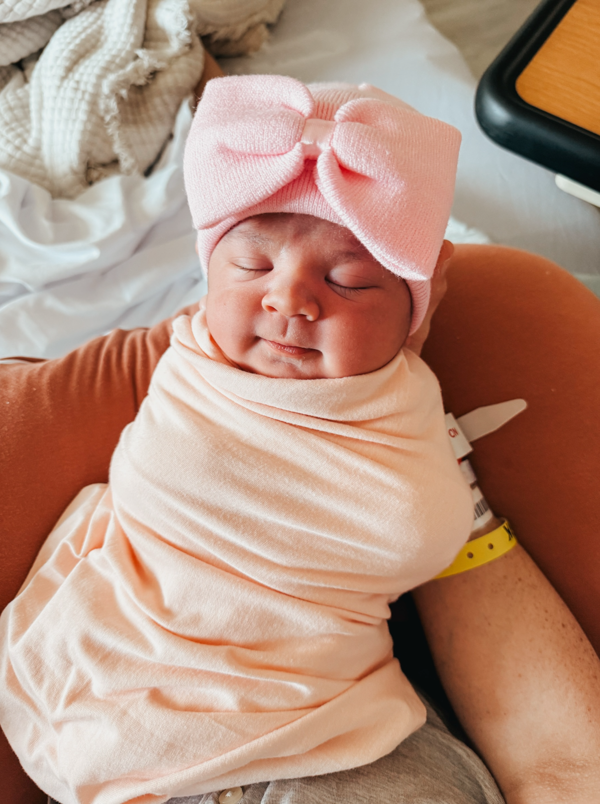 Newborn baby with a pink bow headband, peacefully swaddled, resting in a cozy setting.