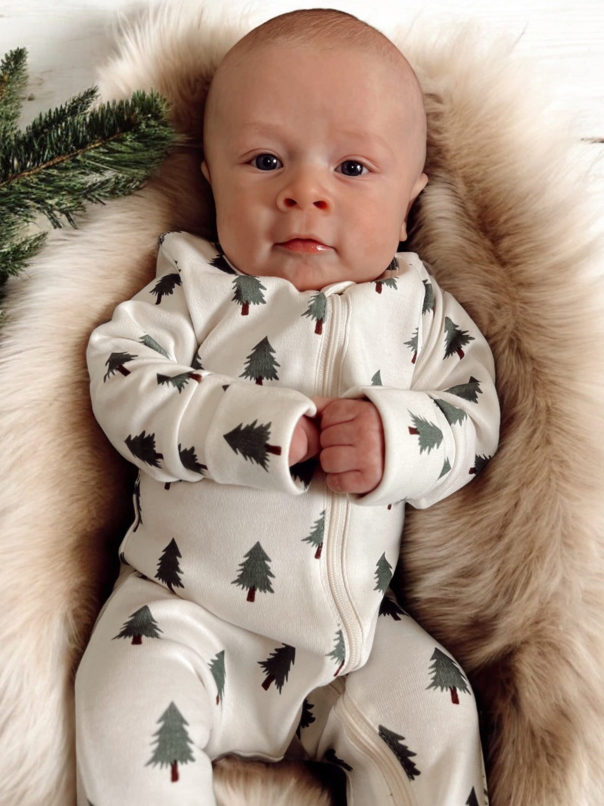 Baby in a tree-patterned onesie, resting on a soft fur blanket with greenery nearby.