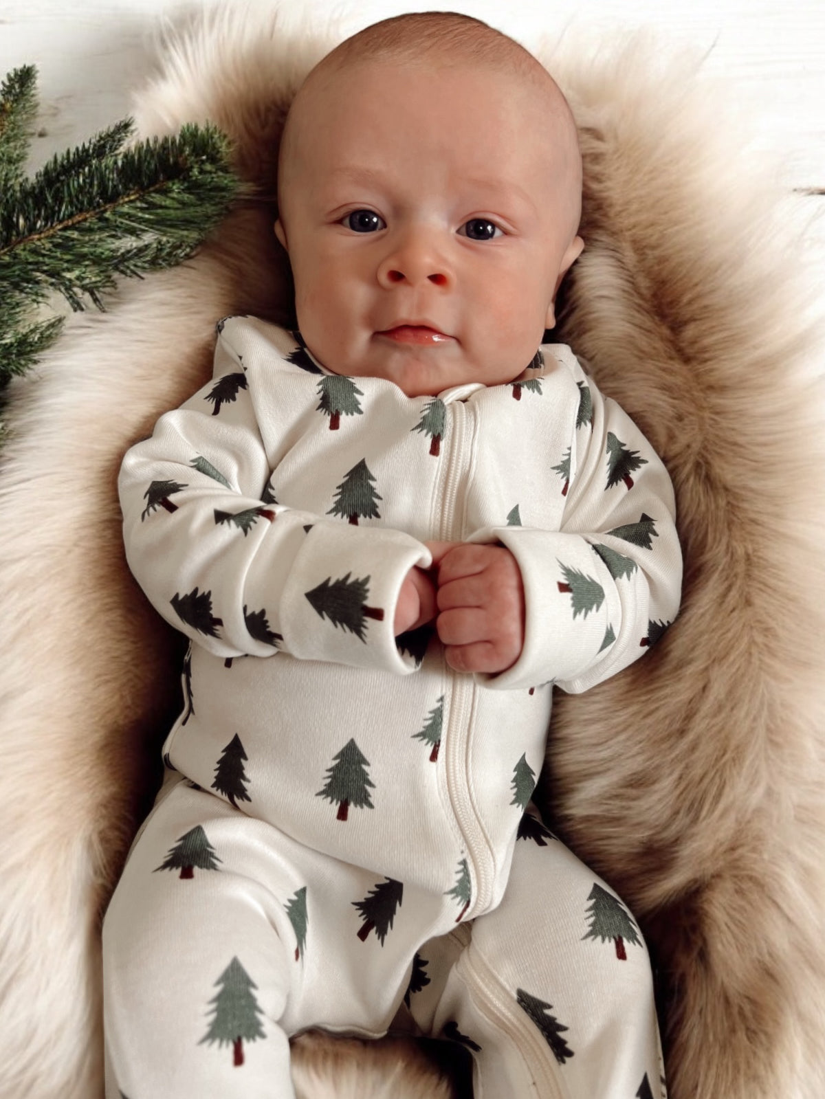 Baby in a tree-patterned onesie, resting on a soft fur blanket with greenery nearby.
