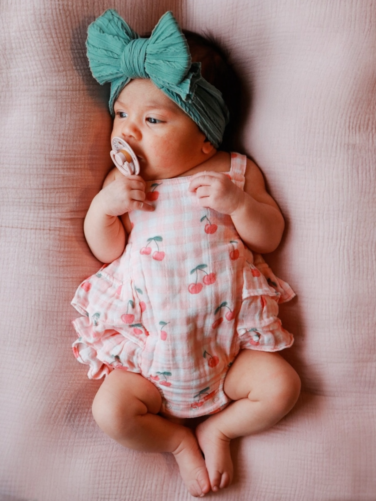 Infant girl in a cherry-patterned onesie with a teal headband, lying on a pink blanket, holding a pacifier.