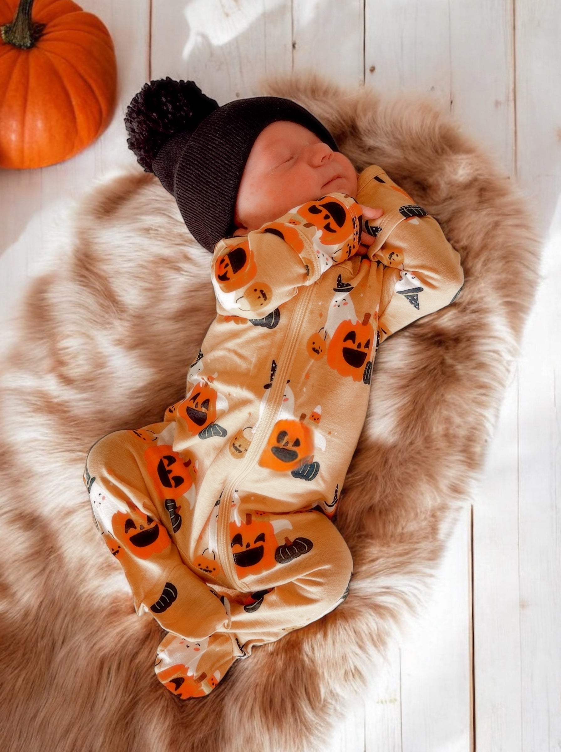 Sleeping baby in a pumpkin-patterned onesie, wearing a black hat, on a fluffy blanket with a pumpkin in the background.