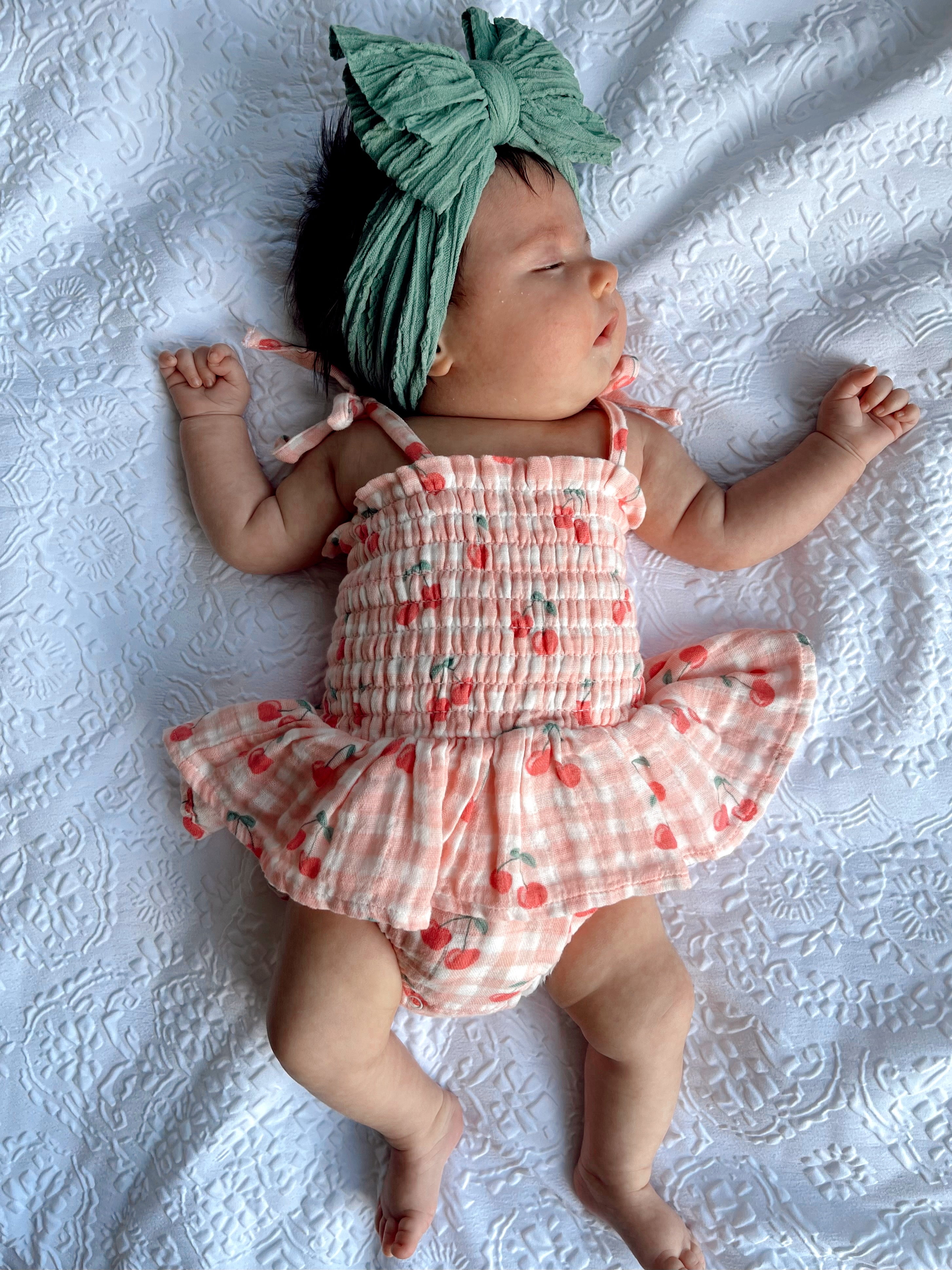 Infant girl in a cherry-patterned outfit and green headband, lying on a white lace background.