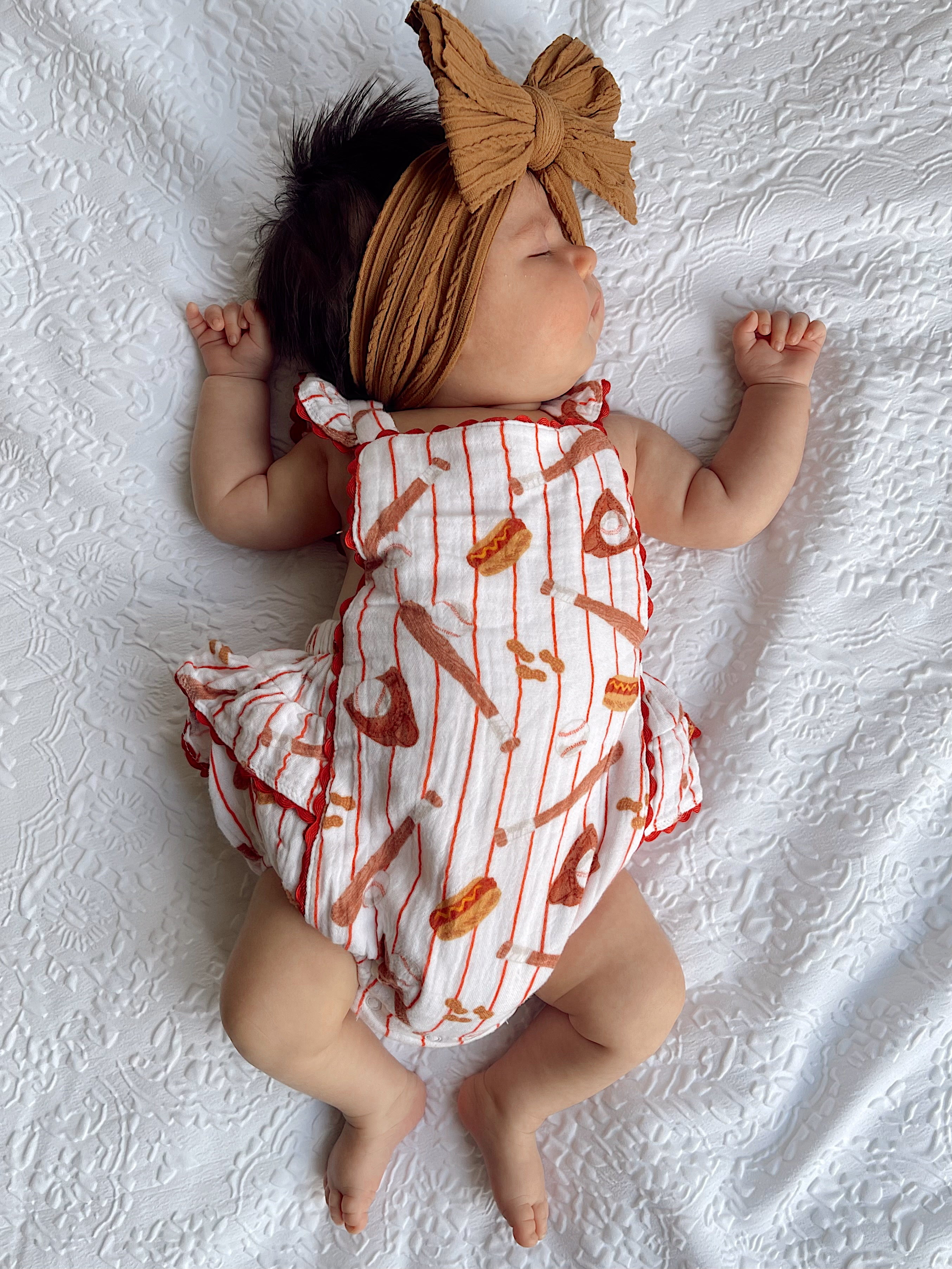 Baby girl sleeping on a white textured blanket, wearing a food-print romper and a large bow headband.