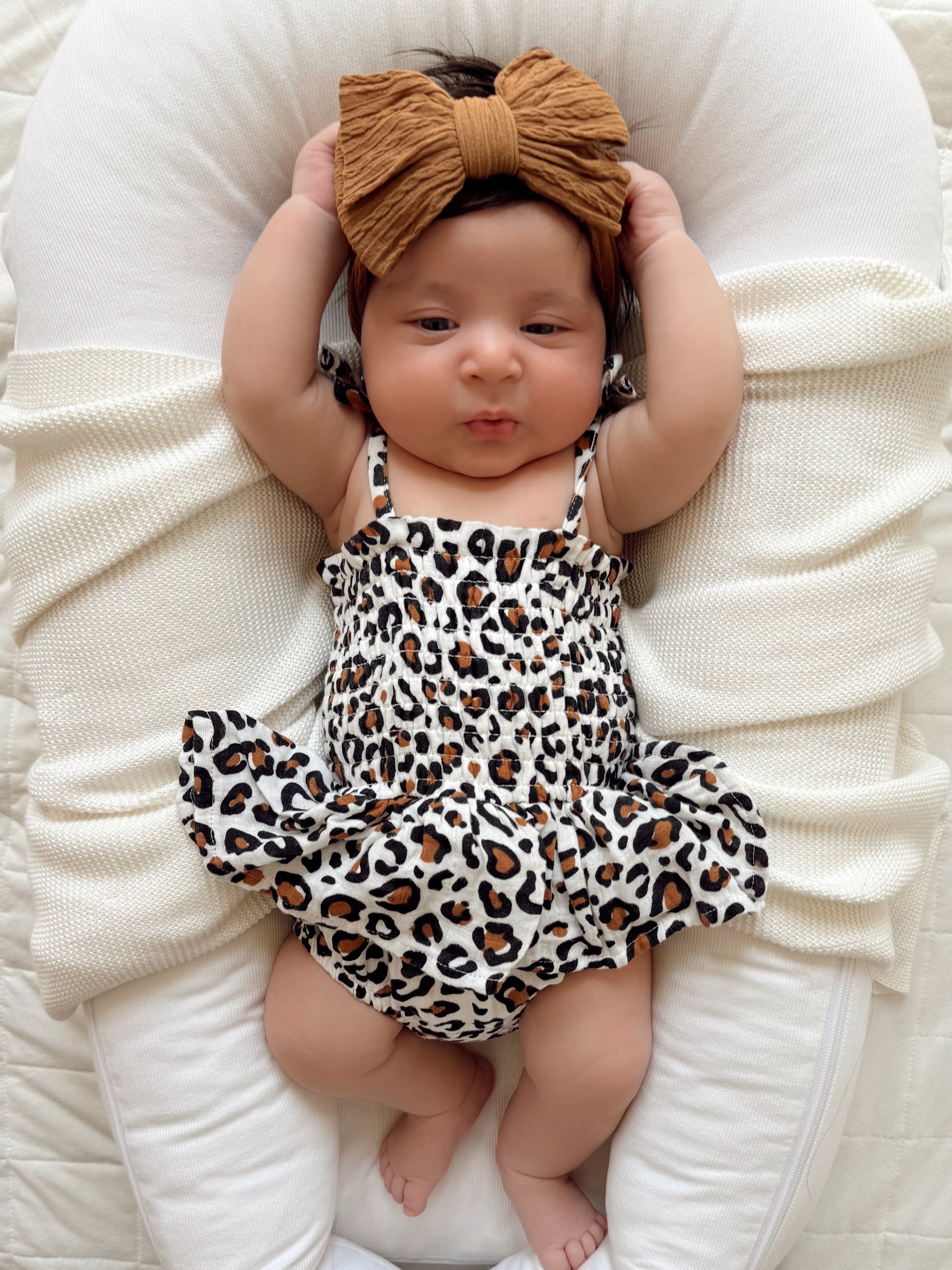 Baby girl in a leopard print dress and a large bow, lying on a cozy white blanket with arms raised.
