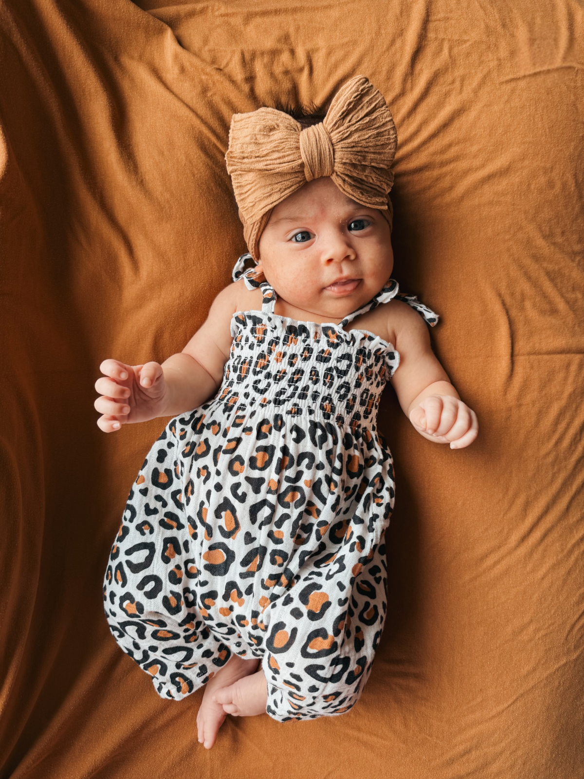 Baby girl with a tan bow headband, wearing a leopard print outfit, lying on an orange blanket.