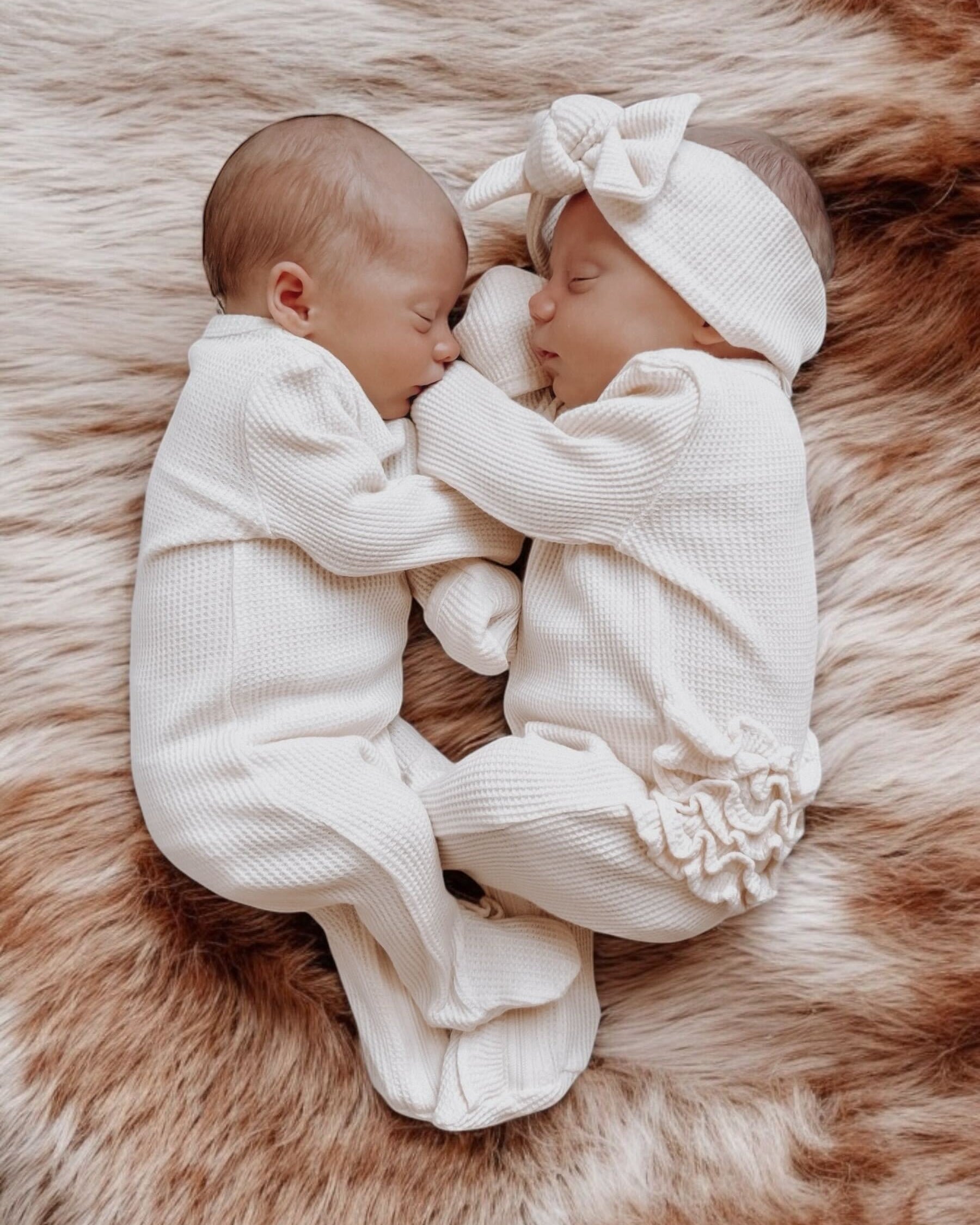 Two newborns in white outfits peacefully sleeping on a soft, textured blanket.