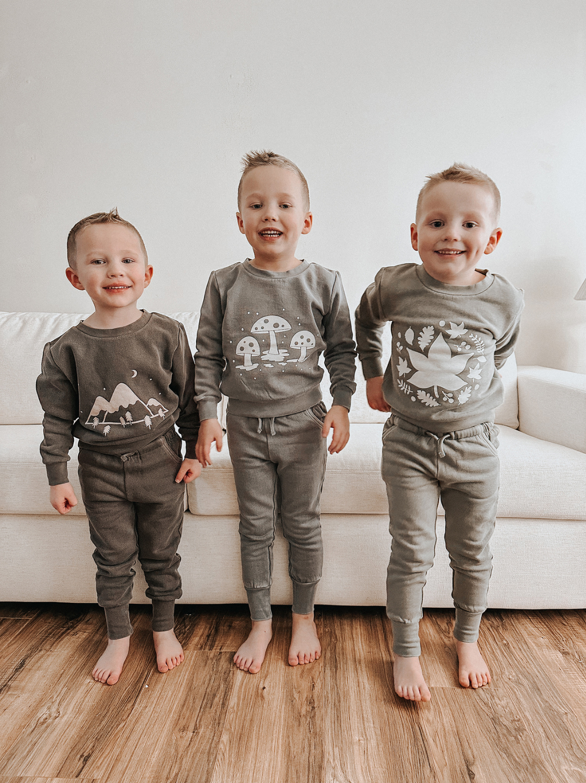 Three smiling boys in matching gray pajamas stand on a wooden floor in front of a white couch.
