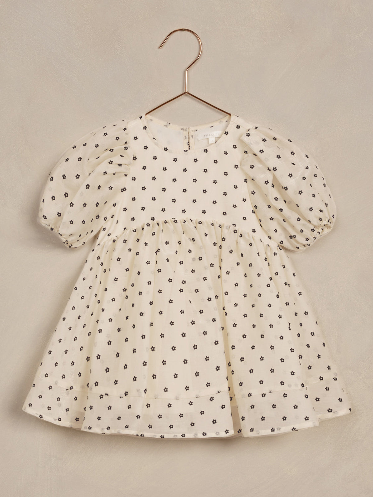 Cream dress with black flower patterns and puff sleeves hanging on a copper hook against a neutral background.