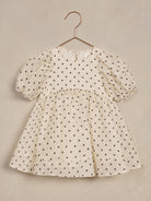 Cream dress with black flower patterns and puff sleeves hanging on a copper hook against a neutral background.