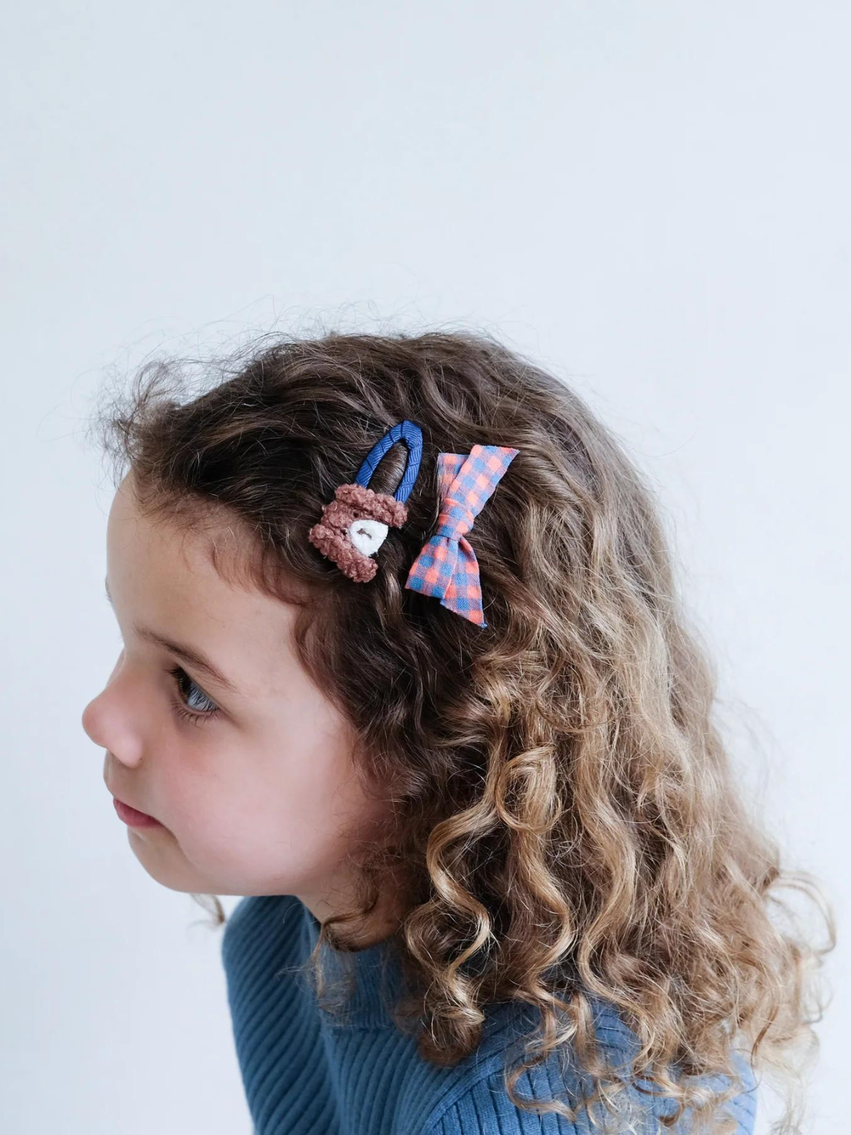 Child with curly hair, wearing a bear and a plaid hair clip, looking to the side against a light background.