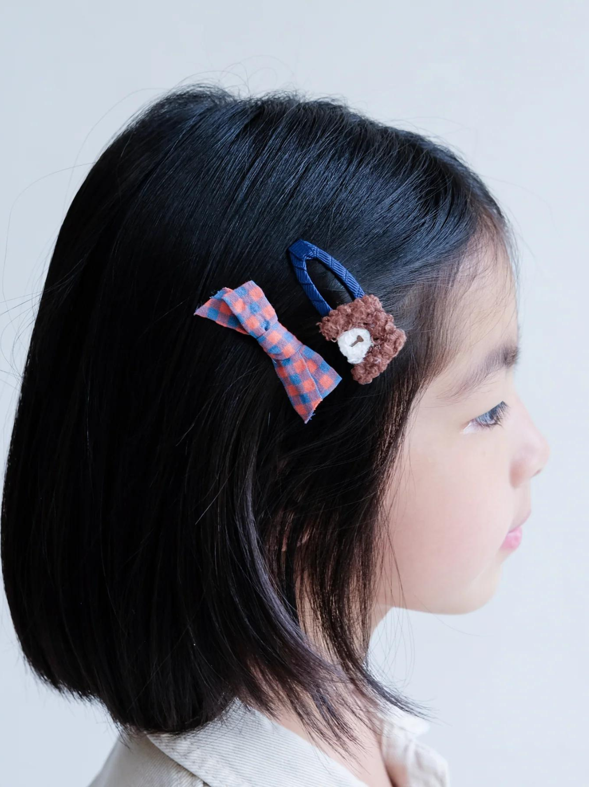 Child with black hair wearing two colorful hair clips: a checkered bow and a bear-shaped clip.