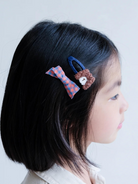 Child with black hair wearing two colorful hair clips: a checkered bow and a bear-shaped clip.