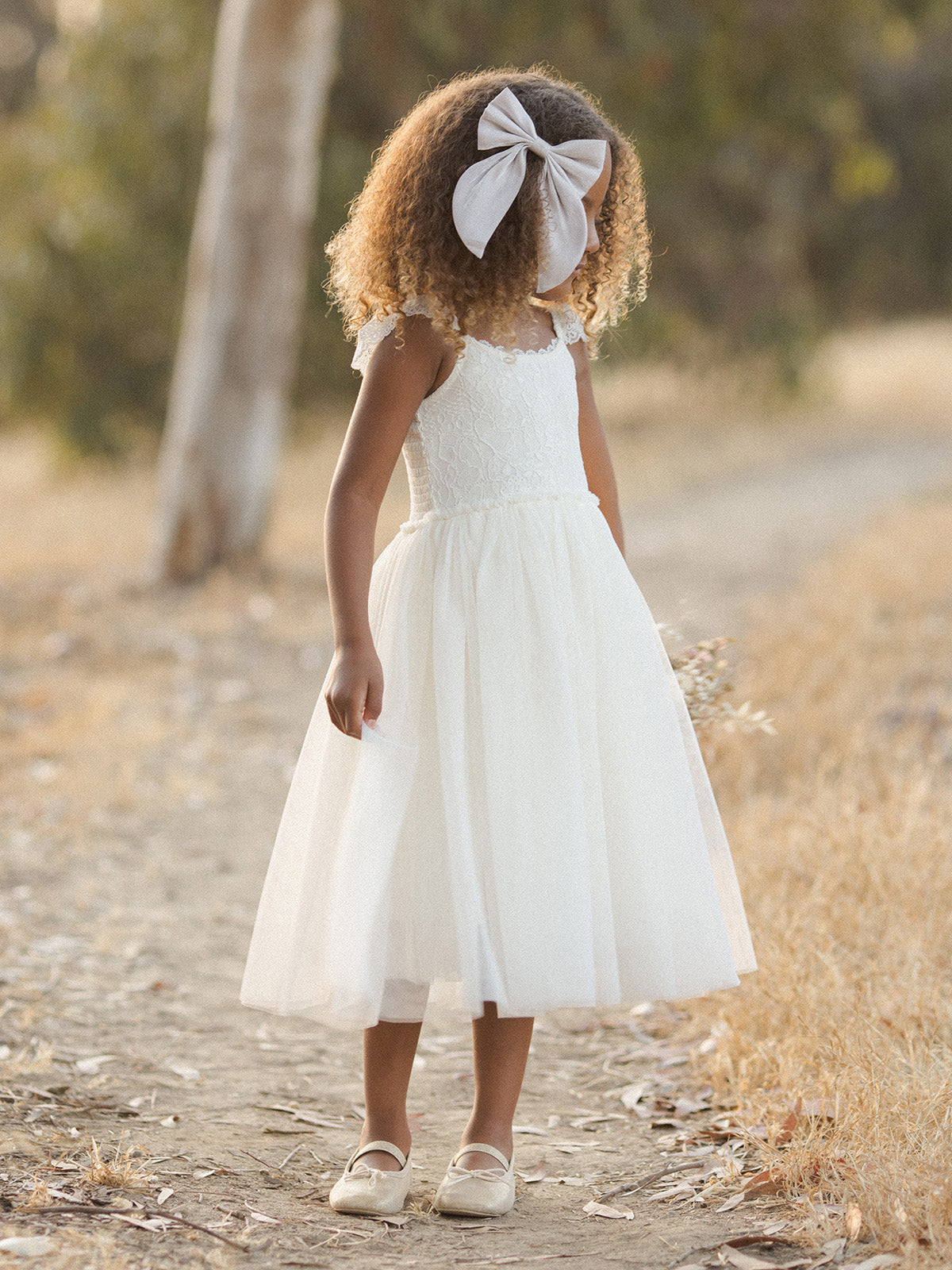 Young girl in a white dress with a large bow, standing on a dirt path surrounded by dry grass and trees.