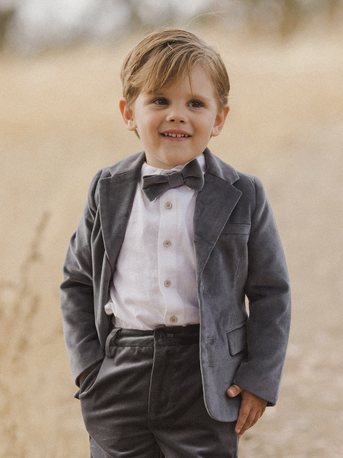 Smiling boy in a gray suit and bow tie standing outdoors, with a blurred natural background.