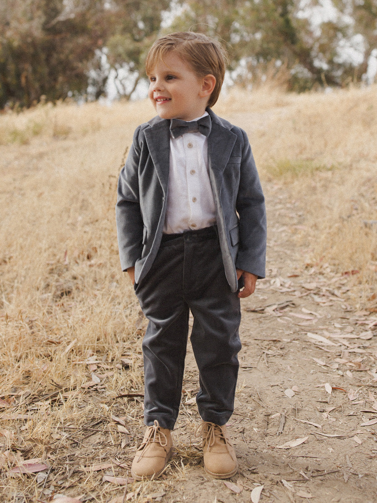 Smiling boy in a suit and bow tie stands on a path surrounded by dry grass and trees.