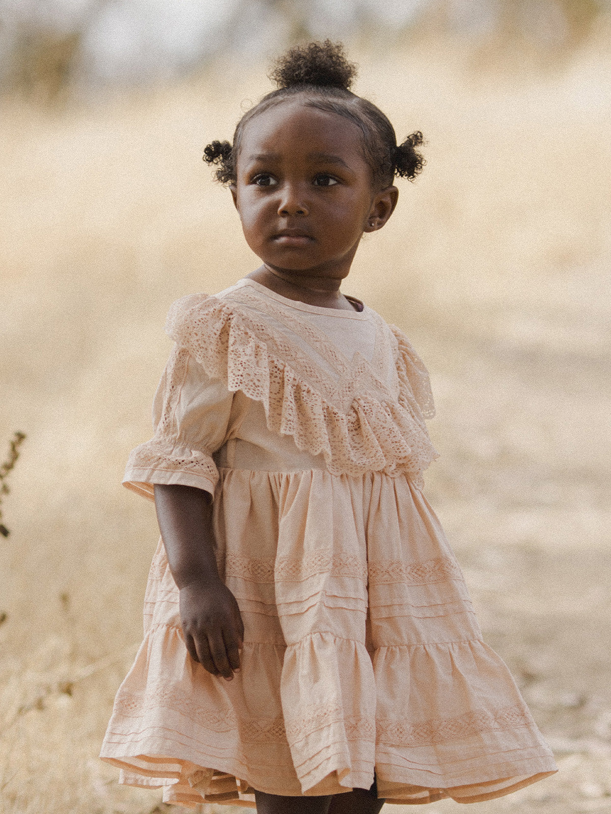 Young girl in a peach dress with lace details, standing outdoors with a thoughtful expression. Background is blurred.