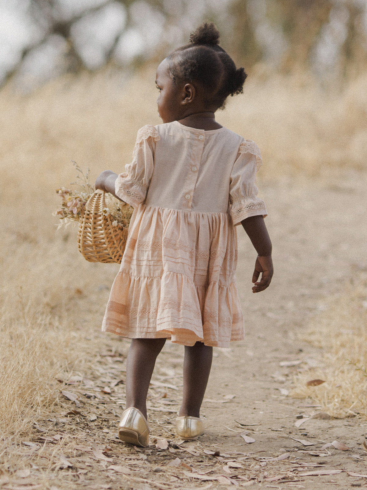 Young girl in a peach dress walking on a path, holding a woven basket with flowers. Natural, soft background.