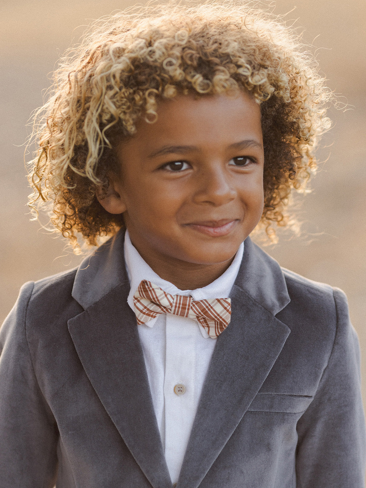 Smiling boy with curly hair wearing a gray suit and striped bow tie against a soft, neutral background.