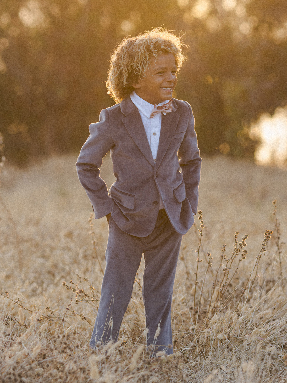 Smiling child in a stylish suit standing in a sunlit field, exuding confidence and joy.