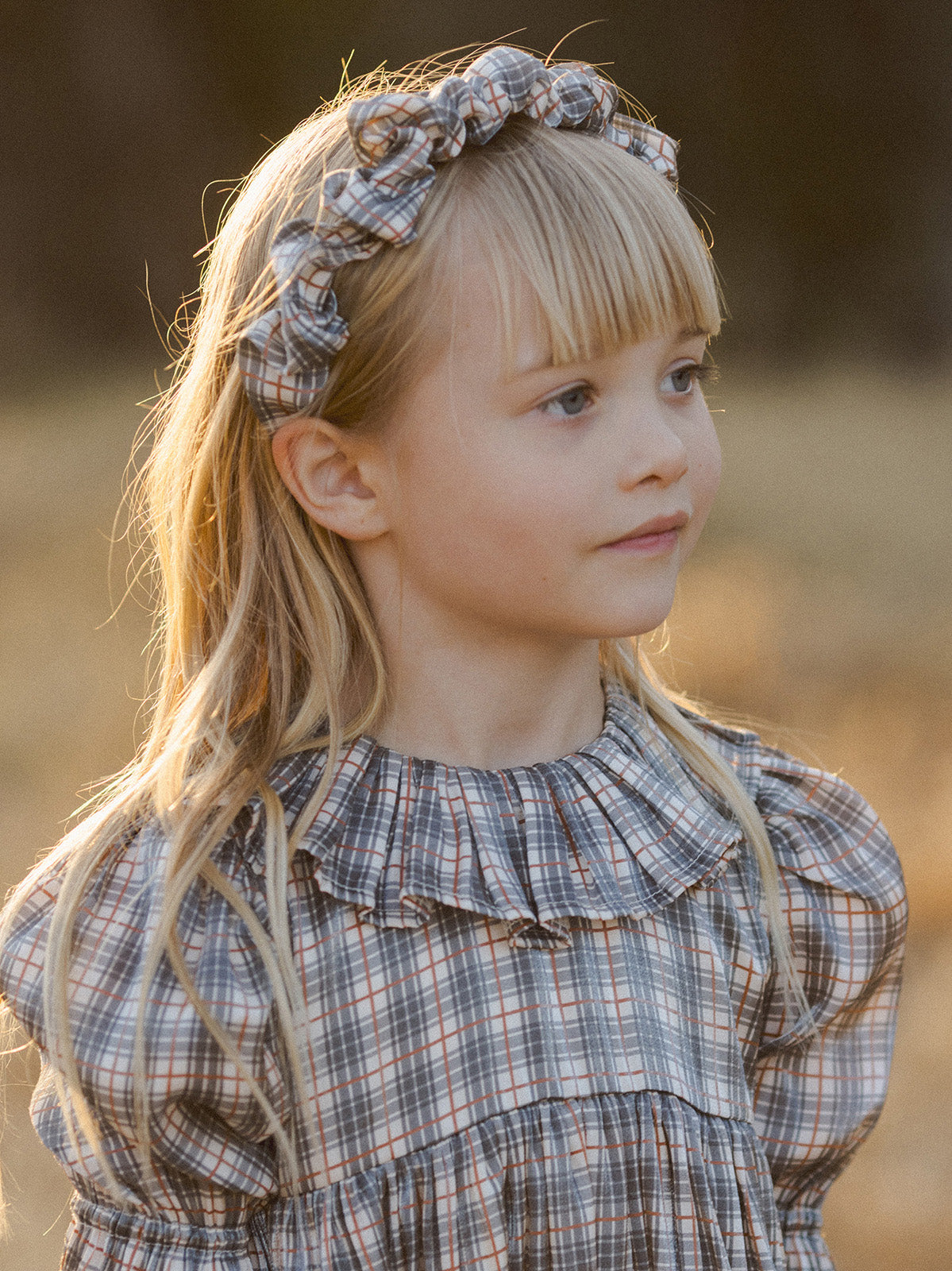 Girl with long blonde hair wearing a checked dress and bow headband, standing outdoors in soft lighting.