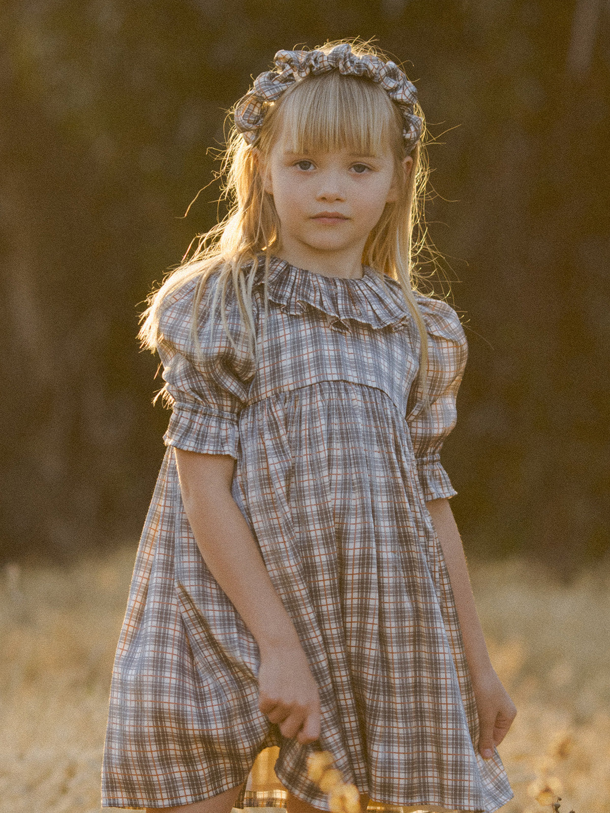 Young girl in a plaid dress and headband stands in a sunlit field, looking thoughtfully at the camera.
