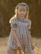 Young girl in a plaid dress and headband stands in a sunlit field, looking thoughtfully at the camera.