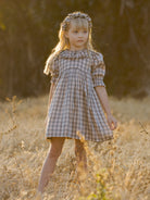 Girl in a plaid dress stands in a sunlit field, with a floral headband and soft, dreamy expression.