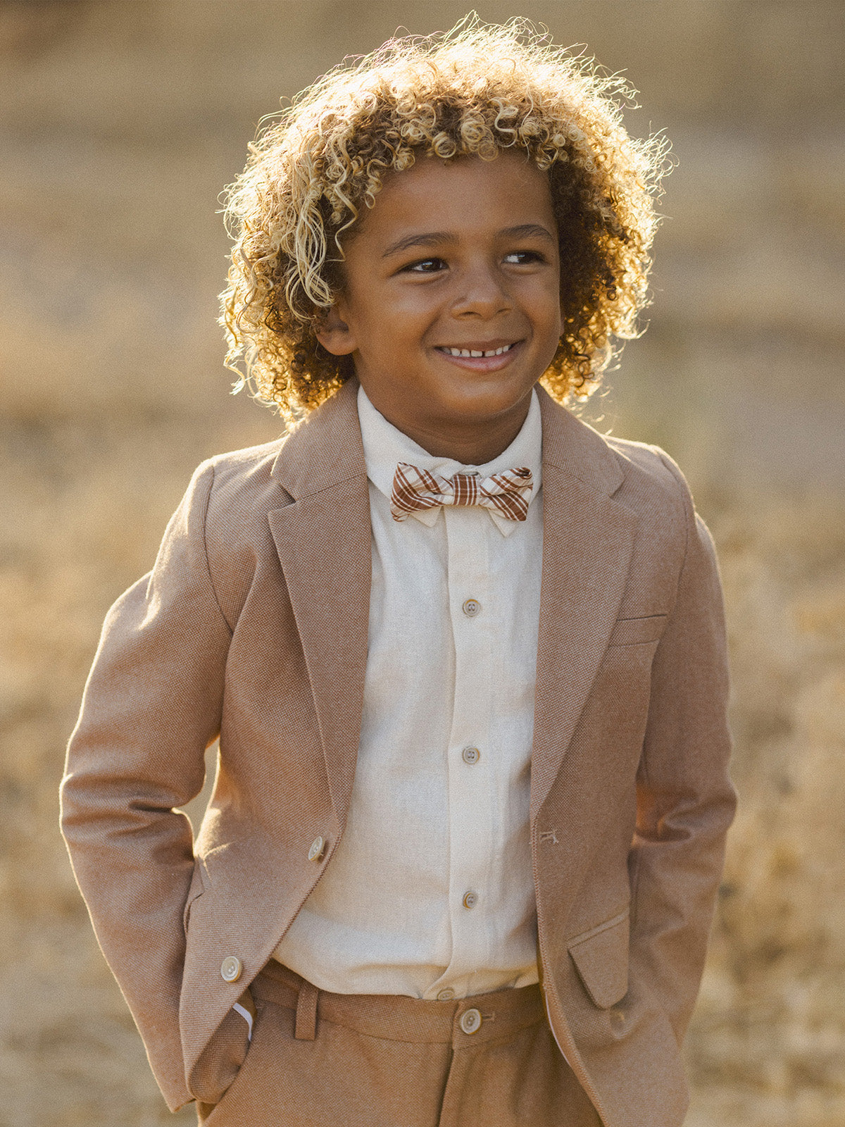Smiling boy with curly hair wearing a tan suit and bow tie, standing outdoors in soft golden light.