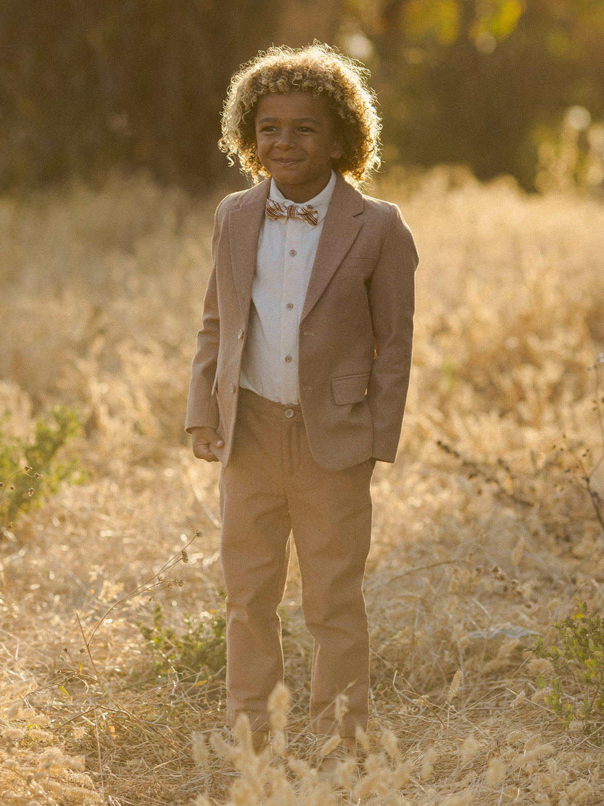 Young boy in a beige suit and bow tie smiles in a sunlit field with tall grass.