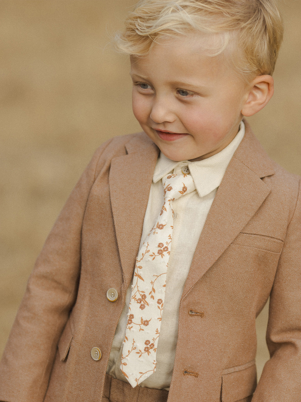 Young boy in a brown suit and floral tie, smiling outdoors against a neutral background.