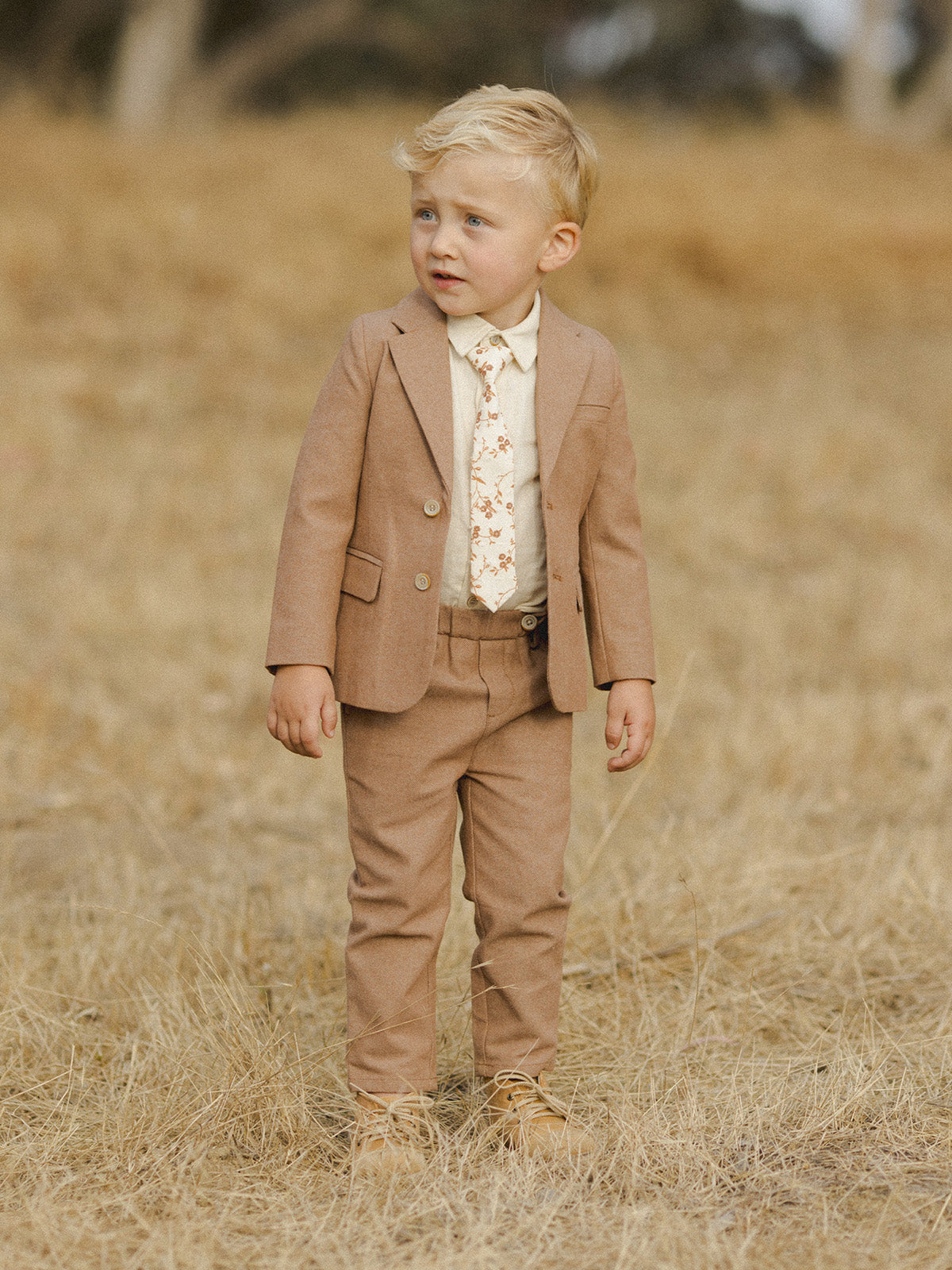 Young boy in a tan suit standing in a field of dried grass, looking off to the side.