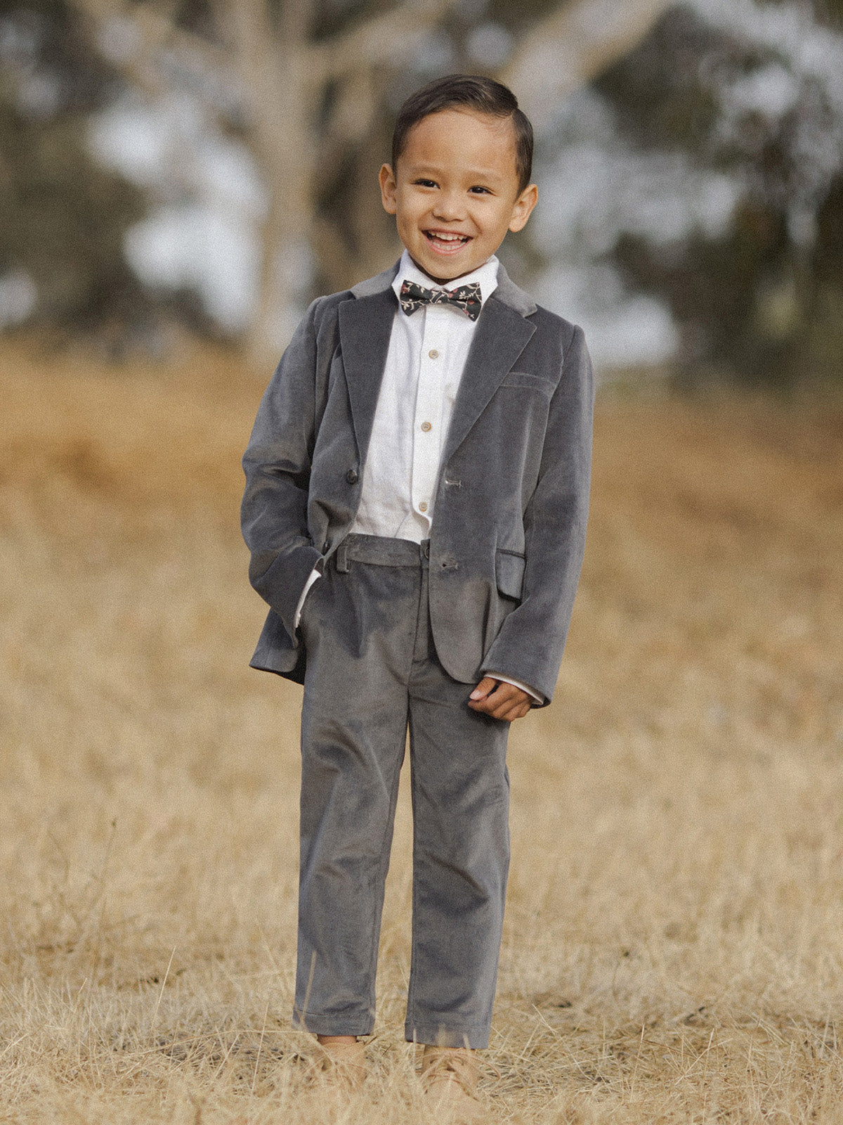 Young boy in a gray suit and bow tie, smiling against a natural background of dry grass and trees.