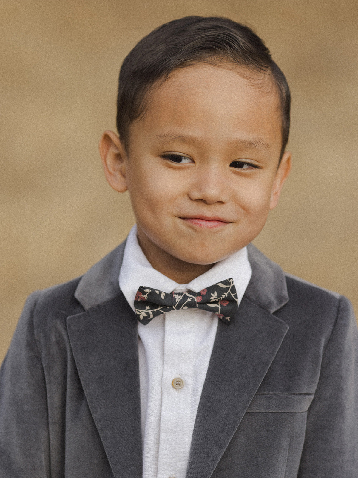 Smiling boy in a gray velvet blazer and floral bow tie, against a soft, neutral background.