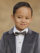 Smiling boy in a gray velvet blazer and floral bow tie, against a soft, neutral background.