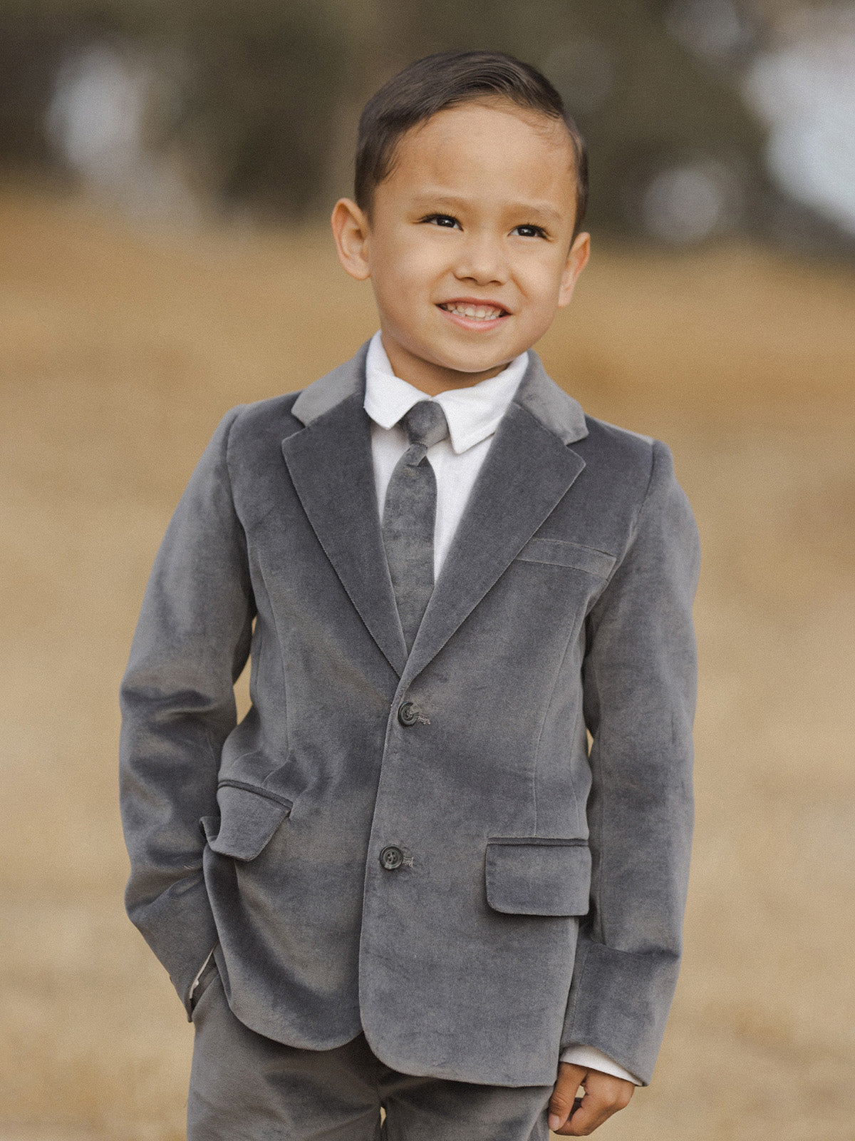 Smiling boy in a gray suit standing outdoors against a blurred natural background.